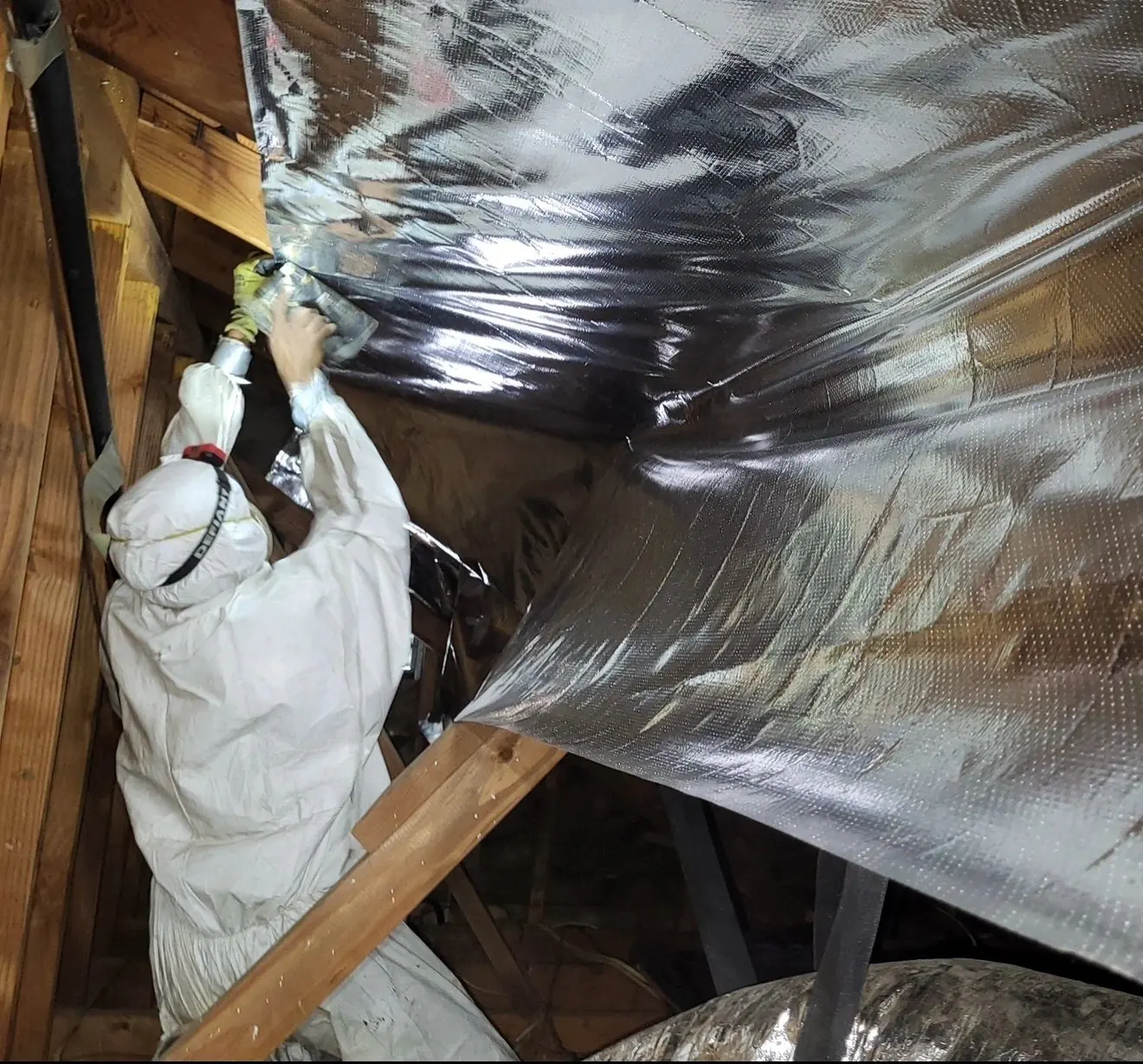Technician in protective coveralls sealing insulated duct in an attic
